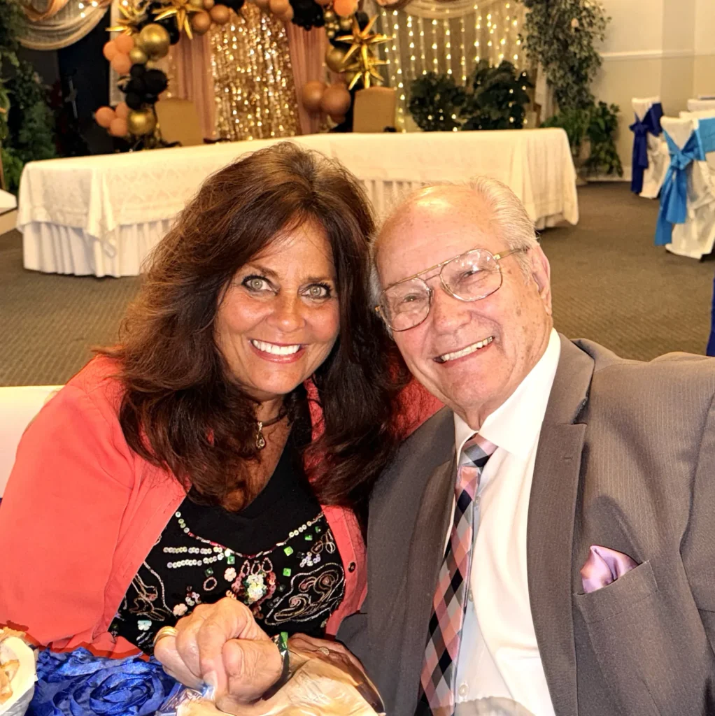 A woman and a man smiling together at a table during a church celebration, with gold and black balloons behind them