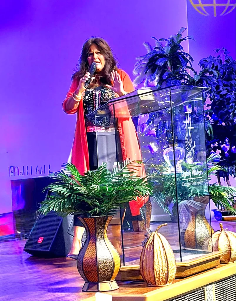 Jennifer Tuttle Gentle speaking with a microphone beside the glass pulpit at Living Waters during the 2026 Homecoming